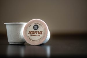 Two Xoma superfoods compostable coffee pods sitting on a table. One is upright, the other is on its side showing the label. The label reads: NEXE Xoma Superfoods. Plant based packaging. Rethink what's possible.