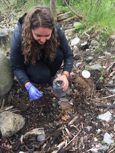 Kennedy Bucci kneels, holding a jar and wearing one purple glove, and picks through microplastics on the shoreline.